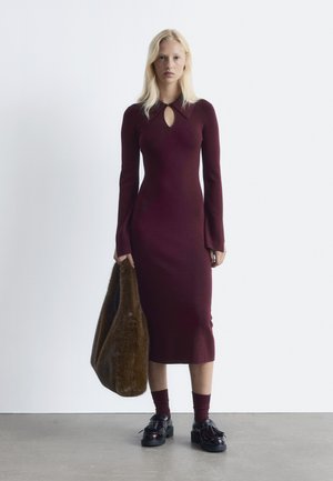 Bordeauxfarbenes Rippkleid mit langen Ärmeln und einem Schlüsselloch-Ausschnitt, kombiniert mit einer braunen Kunstfelltasche und schwarzen Plateau-Schuhen. Glatte Textur.