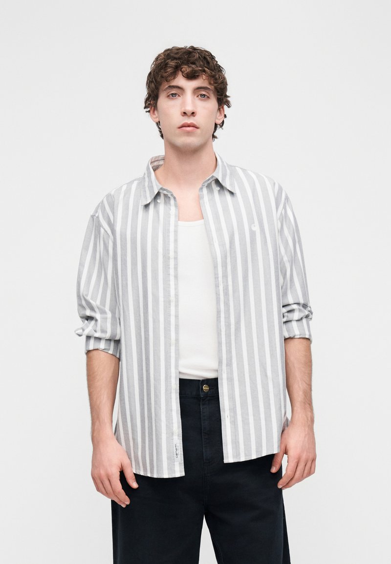 Camicia a righe verticali grigie e bianche con bottoni, tessuto leggero, maniche lunghe arrotolate fino ai gomiti, indossata sopra una canottiera bianca, abbinata a pantaloni scuri.