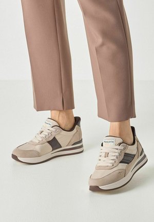 Beige und braune Sneaker, getragen zu hellbraunen kurzen Hosen bei einer Person, die auf einem weißen Boden steht.
