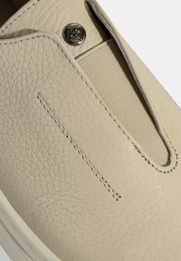 Beige instap schoen van textuur leer, met een canvas elastische inzet en een metalen accentknop. Zichtbare stiksel details aan de zijkant.