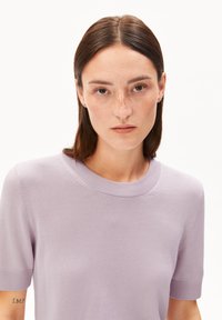 Donna con capelli castani lisci e lentiggini, che indossa un maglione viola a maniche corte, guarda in avanti con un'espressione neutra.