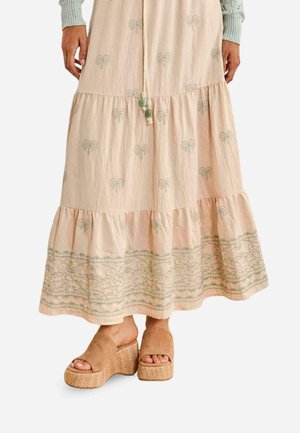 Falda de volantes beige claro con patrones decorativos y palmeras verdes, combinada con sandalias marrones de plataforma y un top de punto gris claro.