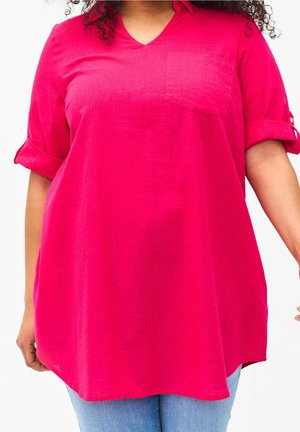 Frau trägt ein leuchtend pinkes langärmliges Hemd mit hochgekrempelten Ärmeln und hellblaue Jeans, steht vor einem schlichten weißen Hintergrund.