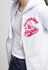 Sudadera gris con cremallera y logotipo bordado en rosa "Superdry Angels" en el lado izquierdo; cuenta con cordones y una textura suave. Llevada sobre una camiseta blanca.