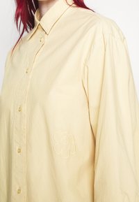 Personne aux cheveux roux portant une chemise ample jaune pâle à boutons avec une fleur brodée discrète sur le côté.