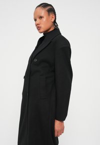 Manteau en laine noir avec une coupe ajustée, des manches longues, deux boutons et des poches latérales, présentant un design classique à revers cranté.