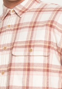 Koszula w kratę zapinana na guziki w odcieniach kremowym, różowym i brązowym. Posiada dwie kieszenie na klatce piersiowej oraz klasyczny kołnierzyk z naturalnymi drewnianymi guzikami.
