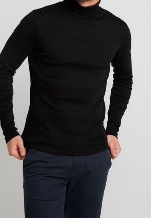 Homme portant un col roulé noir ajusté à manches longues et un pantalon bleu foncé, sur fond clair uni.