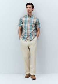 Camicia a maniche corte a quadri in turchese, beige e bianco, con colletto abbottonato. Abbinata a pantaloni beige e scarpe in camoscio marrone.