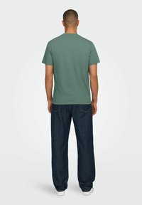 Green cotton t-shirt with a crew neckline, paired with loose-fit dark denim jeans and white sneakers, viewed from the back.
