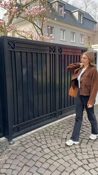 Casaco de camurça castanho com detalhes em botões, t-shirt branca, jeans pretos e ténis brancos. Caminho de calçada e um portão preto ao fundo.