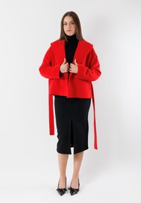 Cappotto rosso accorciato con un colletto oversize e maniche lunghe, indossato sopra un vestito nero aderente con uno spacco anteriore. Tacchi neri a punta.