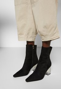 Botas de tobillo de ante negro con puntera afilada y tacón bloque transparente. Presenta una cremallera lateral y una superficie lisa y texturizada.