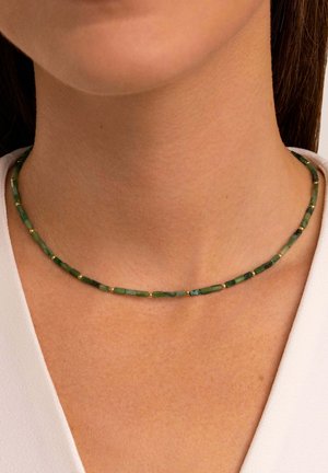 Femme portant un collier vert à perles avec de petites perles séparatrices dorées sur un haut clair, visible du cou à la clavicule.