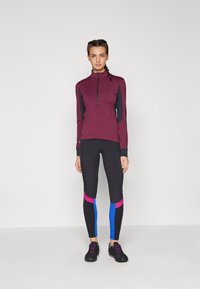Bordeaux színű, fekete részletekkel rendelkező sportfelső, félig cipzárral, fekete leggings-szel párosítva, amelyek rózsaszín és kék panelekből állnak. Fekete lábbeli, lila részletekkel.