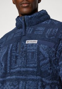 Dunkelblauer Fleece-Pullover mit hohem Kragen und Halb-Reißverschluss. Besitzt eine weiche, strukturierte Oberfläche und eine kleine Brusttasche mit Markenlogo.