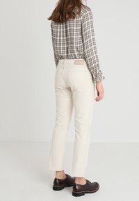 Bílé zkrácené džíny s třepeným lemem, spárované s kostkovanou košilí s dlouhým rukávem. Boty jsou tmavě hnědé s texturovaným povrchem.