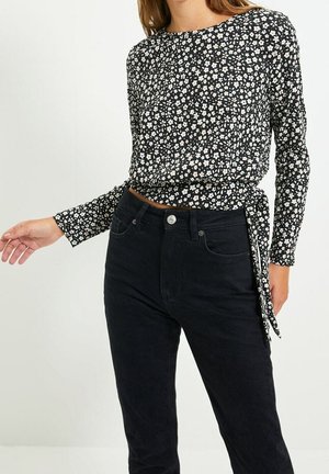 Frau trägt ein schwarzes, langärmeliges, florales Oberteil mit Seitenschleife und hoch taillierte schwarze Jeans vor einem einfarbigen Hintergrund.