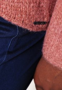 Lyserød strikket trøje med tekstureret finish, kombineret med mørkeblå denimjeans; har et lille sort mærke med brandnavn på trøjen.