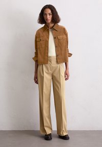 Bruine corduroyjas met twee borstzakken, gecombineerd met beige wijde broek en donkere schoenen. De jas heeft een kraag en knoopsluiting.