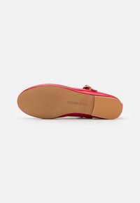 Chaussure en cuir verni rouge avec un bout arrondi, un petit talon et une semelle en caoutchouc texturée. Présente un petit détail de bride avec boucle.