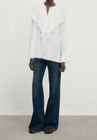 Person iført en hvid, rynket bluse med lange ærmer og mørkeblå, bredbenede jeans, parret med sorte sandaler.