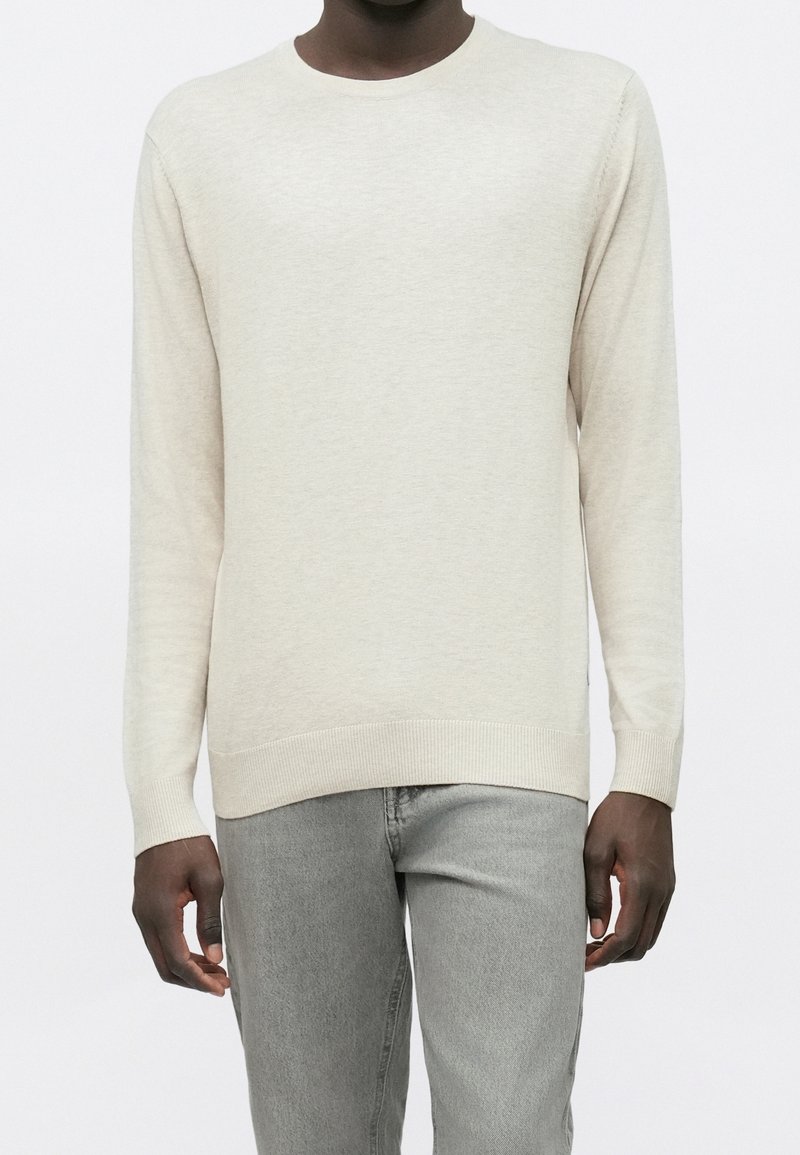 Homme portant un pull beige uni à manches longues et un jean gris clair, debout devant un fond blanc.