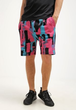 Mann trägt bunte Shorts mit abstraktem Muster in Pink, Blau und Schwarz, kombiniert mit schwarzen Sneakers und einem schwarzen Hemd.