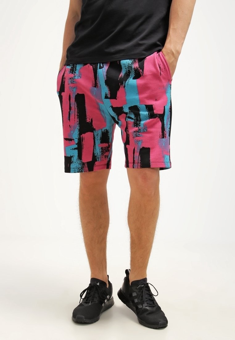 Homme portant un short à imprimé abstrait coloré en rose, bleu et noir, associé à des baskets noires et une chemise noire.