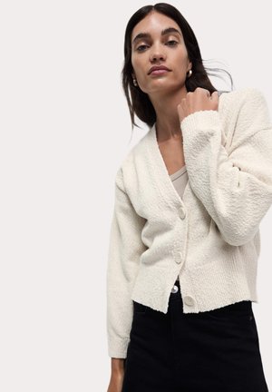 Weißer strukturierter Cardigan mit langen Ärmeln, mit V-Ausschnitt und vorderen Knöpfen, getragen über einem beigen Top und schwarzen Hosen.