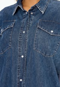 Camicia di denim di colore blu scuro con apertura a bottoni, due tasche sul petto e cuciture a contrasto. Il tessuto ha un aspetto morbido e strutturato.