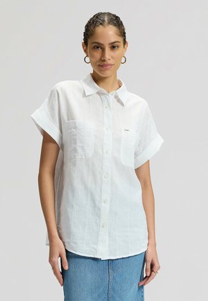 Kobieta ubrana w białą koszulę na krótki rękaw z dwoma przednimi kieszeniami i niebieską spódnicę dżinsową, stojąca na tle jasnego tła.