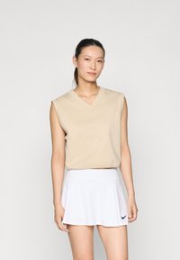 Beige mouwloos topje met een V-hals en een cropped fit, gecombineerd met een witte klokrok met een zwart Nike-logo op de zoom.