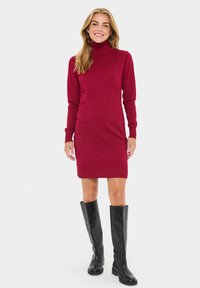 Burgundy poolokauluspaita, jossa on pitkät hihat, tiukka muotoilu ja ribattu yksityiskohtia, yhdistettynä polven ylle ulottuviin mustiin nahkasaappaisiin.