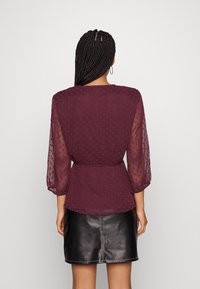 Blusa bordeaux con maniche a sbuffo trasparenti e fantasia a pois, abbinata a una mini gonna in pelle nera. Texture liscia e design aderente.