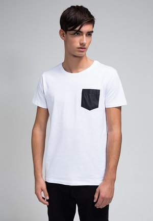 T-shirt bianco in cotone a maniche corte, con una tasca nera sul petto sinistro. Il modello indossa pantaloni neri. Lo sfondo è grigio neutro.