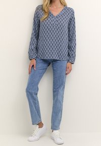 Blouse à manches longues à motifs bleus avec col en V, associée à un jean bleu clair à jambes droites et des baskets blanches. Tissu doux, coupe ample, design géométrique.