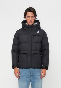 Veste rembourrée noire avec un col montant et une capuche. Elle présente des coutures horizontales, une poche zippée sur la poitrine et un petit logo sur le côté gauche.