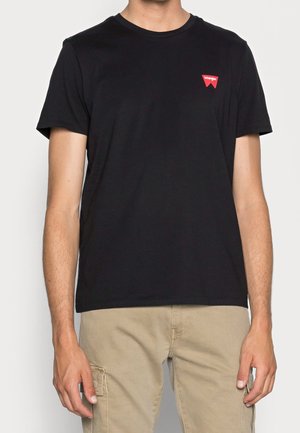 Man draagt een zwart Wrangler t-shirt met een klein rood logo op de borst en beige cargobroek en staat tegen een effen achtergrond.