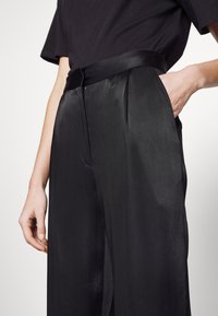 Personne portant un pantalon taille haute en satin noir et un haut noir à manches courtes, avec une main dans la poche du pantalon.