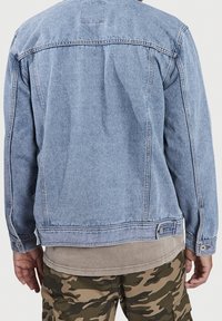 Blauwe spijkerjack met een klassiek ontwerp, zichtbare stiksels, knoopdetails en een relaxte pasvorm, gedragen over een tan kleurig shirt en een gestreepte cargobroek.