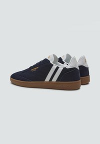 Granatowe zamszowe trampki z białymi paskami i brązową gumową podeszwą. Zawierają złoty tekst logo po bokach i na języku. Teksturalne detale pięty.