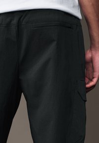 Schwarze Cargo-Hose aus strukturiertem Stoff, mit einer Gesäßtasche und seitlichen Taschen, in einer taillierten Passform und minimalen Nähten.