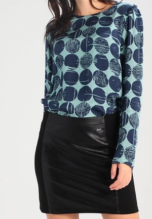 Kobieta w tealowym topie z długim rękawem z granatowymi abstrakcyjnymi kołami i czarnej mini spódnicy o wyglądzie skóry.