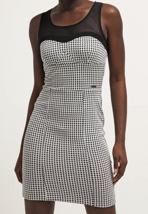 Femme portant une robe cintrée sans manches à motif Pied-de-poule noir et blanc, avec une empiècement transparent noir au niveau du décolleté.
