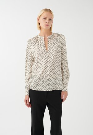 Blusa leggera in crema con motivi circolari marroni scuri, scollatura a V, maniche lunghe e dettaglio arricciato sulle spalle. Abbinata a pantaloni neri.