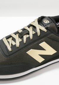 Czarny sneaker z materiałową cholewką, złotymi detalami sznurowadeł oraz dużym złotym logo "N" po boku. Posiada białą podeszwę i teksturowane akcenty.