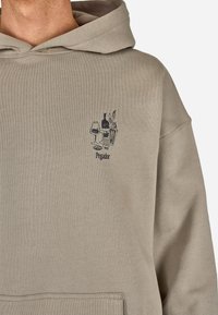 Beige hettegenser med en frontlomme, som har en svart illustrasjon av briller, en flaske og en plante. Mykt materiale med en avslappet passform.