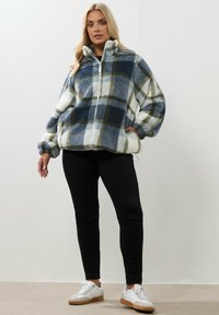 Kvinna klädd i en blå, vit och grön rutig fleecejacka, svarta byxor och vita sneakers, stående mot en enkel vit vägg.