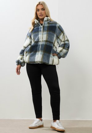 Vrouw in een blauwe, witte en groene geruite fleecejas, zwarte broek en witte sneakers, staand tegen een effen witte muur.
